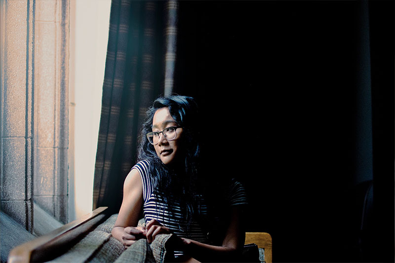Woman with dark hair and glasses in a darkened room staring out a window