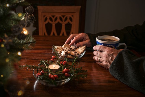 Hands of an elderly single man sitting alone at a table with Christmas cookies, coffee and festive decoration next to an empty chair, lonely holidays after a loss