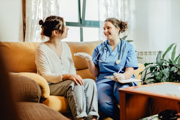 Dark haired female nurse discussing medicare with a dark haired female on a dark yellow couch